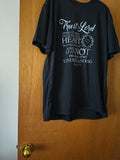 A black short-sleeve graphic t-shirt hanging on a wooden door, featuring a white printed religious quote on the front.
